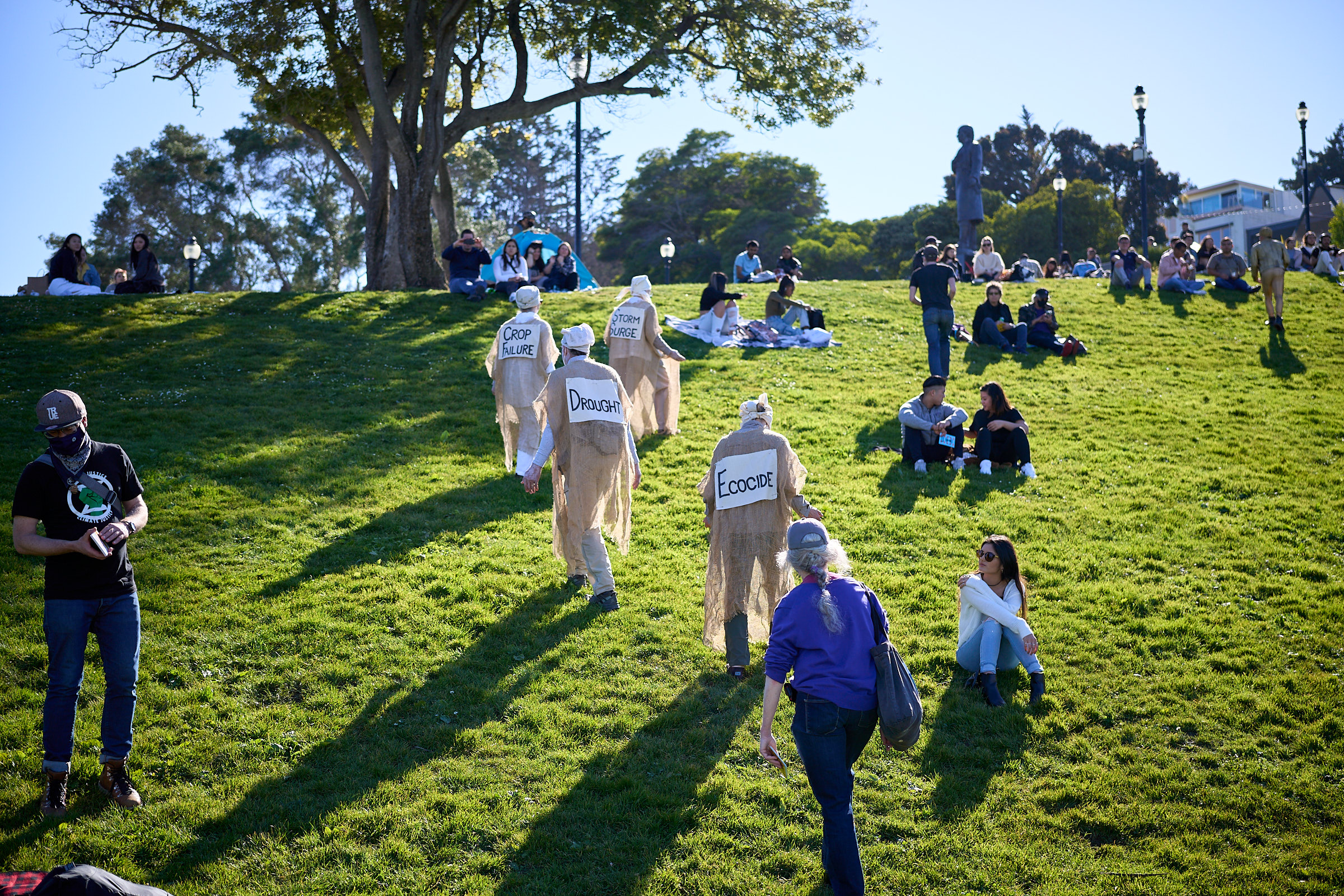 Lamentors dressed in burlap walk among park visitors, with the words "drought", "ecocide" and "crop failure" written on their backs. The green grass is bright from a sunny sky.